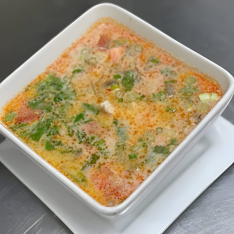 Close-up of a square white bowl filled with creamy Thai soup, garnished with green herbs.