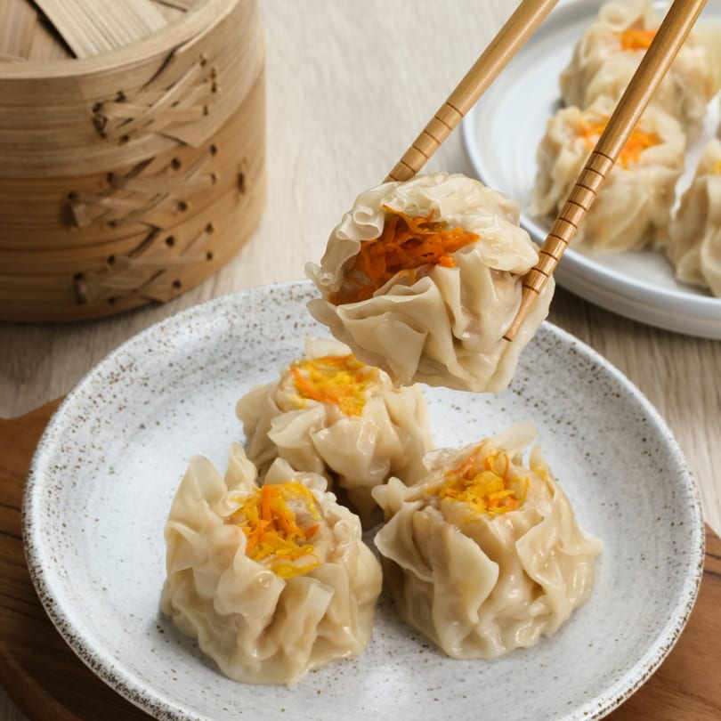 Chopsticks holding a Thai dumpling with orange garnish over a plate of more dumplings.