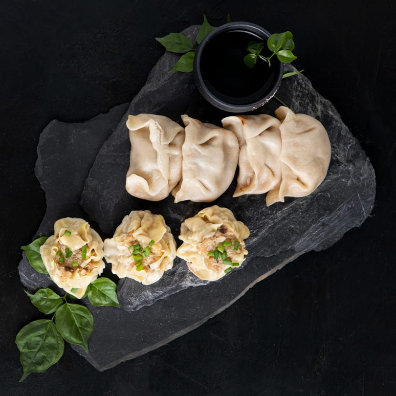 Thai dumplings and shumai with a small bowl of soy sauce on a dark slate board.