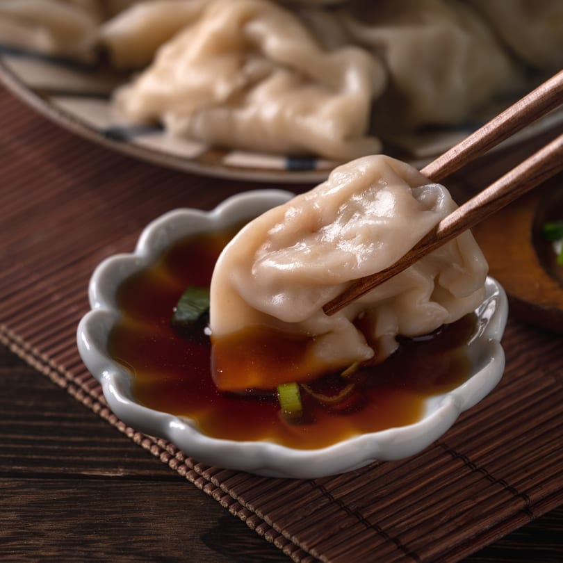 Chopsticks dipping a Thai dumpling into a scalloped white bowl of soy sauce with scallions.