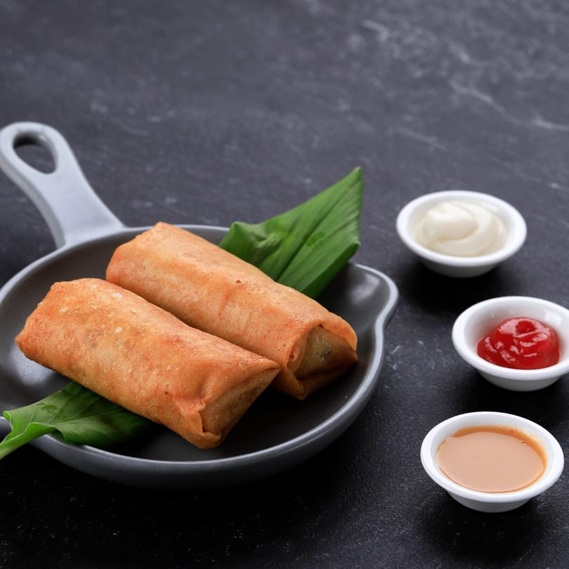 Two crispy Thai spring rolls on a dark plate with green leaves and three dipping sauces.